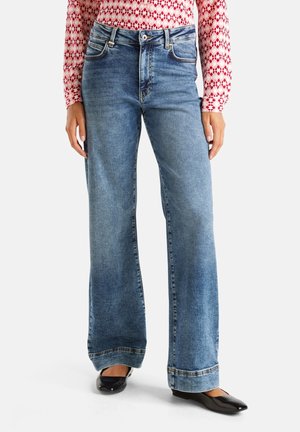 Femme portant un jean bleu à jambes larges, un haut à manches longues rouge à motifs, et des ballerines noires pointues, debout devant un fond blanc.