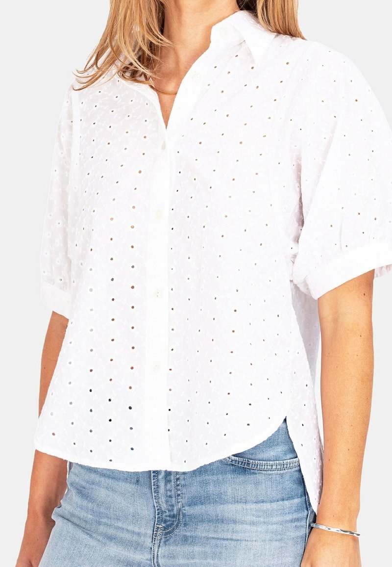 Camicia bianca in cotone a maniche corte con chiusura a bottoni, caratterizzata da ritagli a occhio e una vestibilità rilassata. Indossata con jeans azzurri chiari.