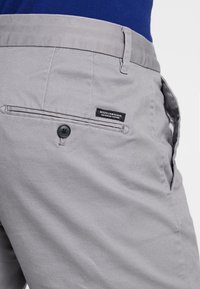 Pantaloni in cotone grigi con chiusura a bottone, tasca laterale e etichetta con marchio. Texture liscia e vestibilità su misura.
