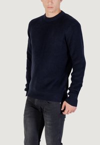 Maglione a coste blu navy con scollo rotondo, maniche lunghe e tessuto testurizzato. Presenta uno spacco laterale con un'etichetta nera a contrasto.