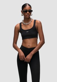 Zwarte geribbelde cropped tanktop met een klein logo, gecombineerd met hoge zwarte leggings. Model draagt oversized zwarte zonnebril en een zilveren ketting.