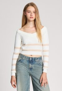 Maglione corto a righe bianche e beige con scollatura scesa sulle spalle, polsini a coste e una texture morbida. Indossato con jeans a vita alta.
