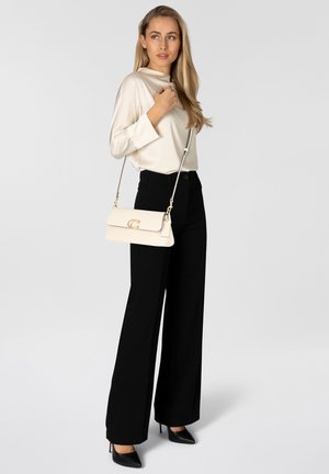 Žena v krémovej saténovej blúzke a čiernych širokých nohaviciach, nosiaca čierne podpätky a nesúca krémovú kabelku cez rameno so zlatými kovaniami.