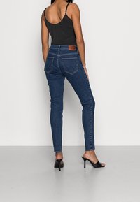 Jeans de mezclilla ajustados en azul oscuro con una cintura de altura media, bolsillos traseros y un parche de cuero marrón en la cintura. Combinados con una camiseta sin mangas negra y tacones.