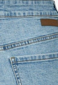 Ljusblå denimjeans med en slät yta, vinklade bakfickor och en rektangulär brun läderlapp ovanför den högra fickan.