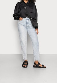 Camicia nera in satin a maniche lunghe, jeans azzurri a vita alta, sandali neri con cinturini, in piedi su un pavimento grigio chiaro contro uno sfondo semplice.