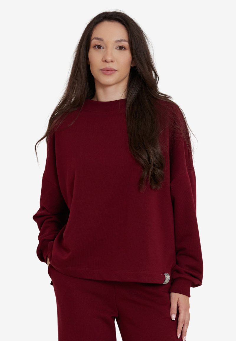 Bordowy sweatshirt z długim rękawem, o prążkowanej strukturze i luźnym kroju. Posiada małą naszywkę z logo blisko krawędzi.
