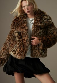 Lederjacke aus Kunstfell mit Leopardenmuster, breitem Kragen und oversized Schnitt. Begleitet von einem grauen T-Shirt und einem schwarzen Rock, die strukturierte Materialien präsentieren.