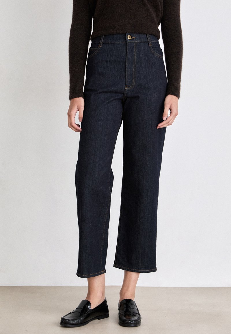 High-waisted dark blue jeans with a straight leg cut, contrasting stitching, and a button closure, paired with black loafers.
