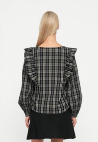 Chemise à carreaux noir et gris avec des volants le long des épaules, manches longues et ourlet arrondi, associée à une jupe noire.