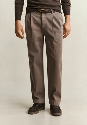 Man wearing taupe straight-leg trousers, dark brown braided belt, dark brown sweater, and dark brown sneakers standing on a light floor.