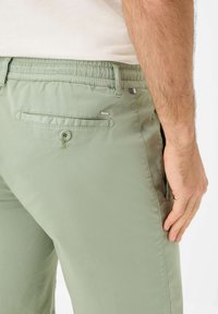 Pantaloni verde chiaro con una texture liscia, dotati di una vita elastica, tasche laterali e una tasca sul retro con bottone. Etichetta con logo visibile.