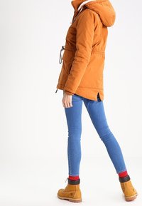 Orange isolerad jacka med huva, dragsko i midjan och texturerad foder, kombinerad med blå tajta jeans och bruna ankelskor.