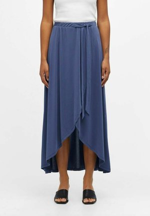 Kvinde iført en blå midilang nederdel i wrap-stil med bindebånd foran og asymmetrisk kant, kombineret med en hvid ærmeløs top og sorte åbne sandaletter.