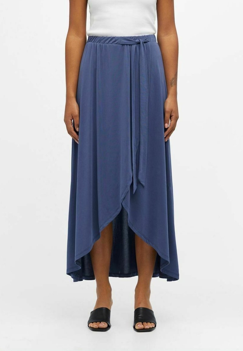 Femme portant une jupe midi bleue style portefeuille avec un nœud devant et un ourlet asymétrique, associée à un top blanc sans manches et des sandales noires à bout ouvert.