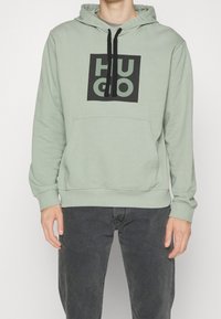 Zelená mikina s kapucí a černým čtvercovým grafickým prvkem s nápisem "HUGO" ve světle zelené. Má přední kapsu a černé šňůrky na kapuci.
