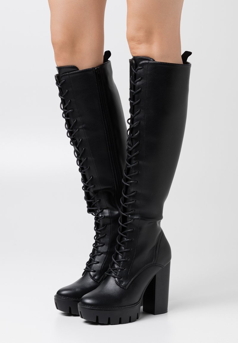 Bottes hautes en cuir noir à lacets avec talons hauts épais et fermetures éclair latérales portées sur des jambes nues sur un fond blanc.