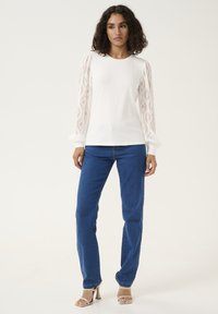 White long-sleeve top with lace details on sleeves, paired with blue jeans and beige heels. The top has a round neckline and a fitted silhouette.