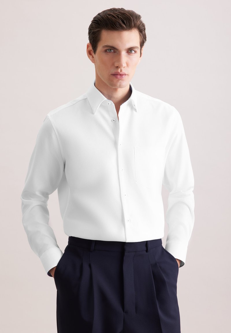 Jeune homme aux cheveux foncés portant une chemise blanche impeccable boutonnée et un pantalon bleu marine plissé, les mains dans les poches, fond neutre.