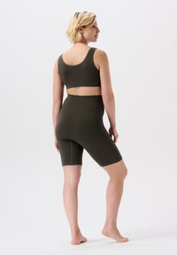 Kvinde står barfodet iført en ribbede olivenfarvet sports-bh og højtaljede cykelshorts, med ryggen til og hovedet vendt til siden.