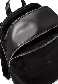 Zaino in pelle nera con finitura lucida, chiusura con zip, rivestimento interno in tessuto e tasca anteriore con pattina. Logo visibile sul davanti.
