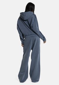 Femeie cu păr lung și închis la culoare, purtând un hanorac larg cu glugă de culoare gri închis și pantaloni evazați, stând cu fața spre depărtare pe un fundal alb.