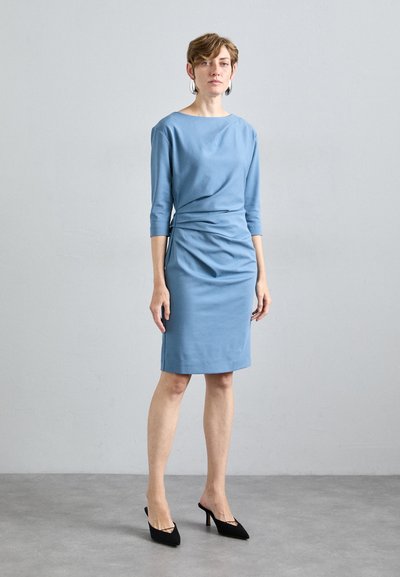 Vestido azul ajustado con escote barco y mangas de tres cuartos. Textura suave y detalle fruncido en el lateral. Combinado con tacones altos negros.