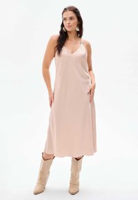 Nudefarbenes Midi-Kleid mit V-Ausschnitt, dünnen Trägern und glattem, satinähnlichem Stoff; kombiniert mit beigen spitzen Stiefeln.