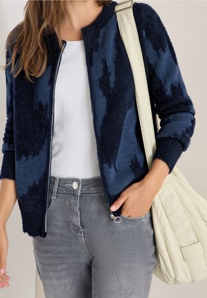 Woman wearing a navy blue patterned zip-up cardigan, white t-shirt, gray jeans, and carrying a cream shoulder bag.