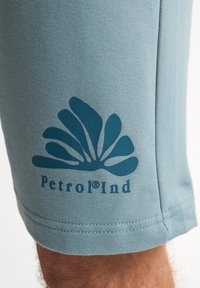 Lichtblauwe shorts van zachte stof hebben een donkerblauw logoontwerp met een gestileerd patroon en de tekst "Petrol Ind" aan de zoom.