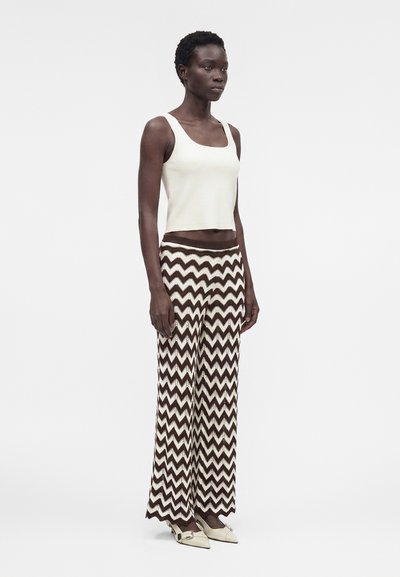 Γυναίκα που φοράει λευκό αμάνικο τοπ και καφέ-λευκό παντελόνι με ψηλή μέση και ζιγκ-ζαγκ σχέδιο, φαρδιά μπατζάκια και λευκά μυτερά παπούτσια.