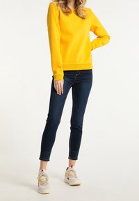 Žlutá mikina s kulatým výstřihem, kombinovaná s tmavými skinny džínami a světlými teniskami s vícebarevnými tkaničkami a detaily.