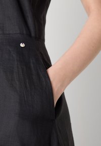 Main droite insérée dans la poche latérale d'une robe en lin noire avec un petit détail de bouton métallique rond.