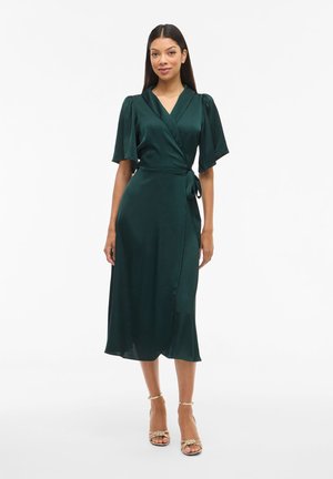 Frau trägt ein dunkelgrünes Satin-Wickel-Midikleid mit kurzen, flatternden Ärmeln und goldenen, hochhackigen Riemchensandaletten, steht vor weißem Hintergrund.