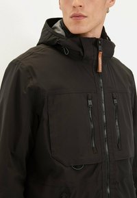 Veste noire résistante aux intempéries avec capuche, dotée de poches zippées, fermeture éclair à l'avant et accents de couture détaillés.
