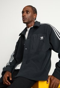 Černá fleecová košile s bundou, která má bílé vertikální pruhy na rukávech, patentky a náprsní kapsu s logem Adidas.