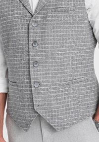 Chaleco gris a cuadros con una tela texturizada, que cuenta con cinco botones, dos bolsillos y un diseño de dobladillo puntiagudo. Camisa blanca visible por dentro.