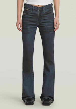 Femme portant un jean évasé bleu foncé et des chaussures noires à bout carré, debout devant un fond clair uni.