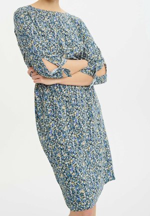 Vrouw draagt een blauw bloemenknielang jurk met 3/4 mouwen met vooraan strikjes en uitsnijdingen. Armen zijn gekruist ter hoogte van de taille.