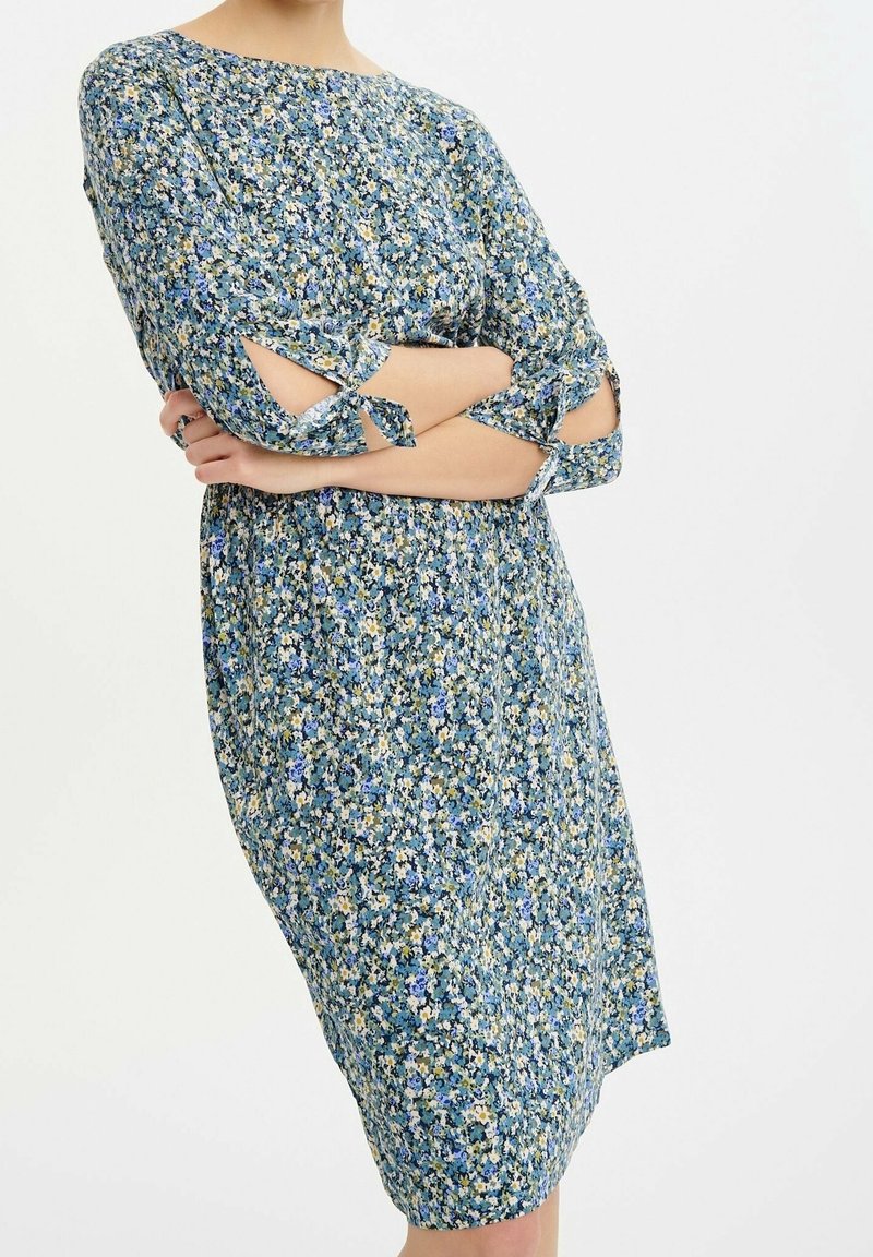 Femme portant une robe bleue à fleurs longueur genou avec des manches 3/4 qui ont des détails de découpes nouées à l'avant. Les bras sont croisés à la taille.