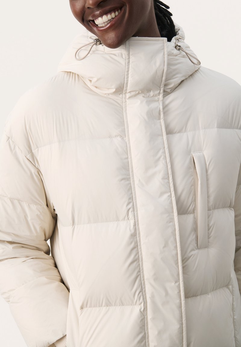 Beżowa kurtka puchowa z kapturem, z zamkiem błyskawicznym z przodu, poziomym pikowaniem i kieszenią boczną. Gładki, lekki materiał.