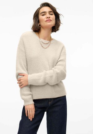 Light beige knitted sweater with long sleeves, ribbed cuffs, and a relaxed fit. Paired with dark blue denim jeans. Simple, textured design.