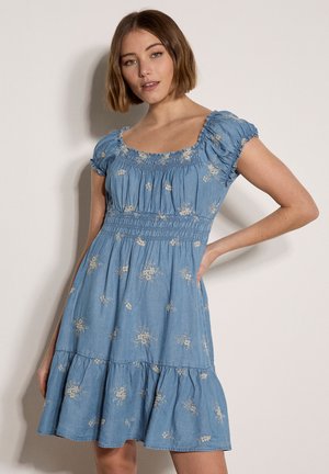 Jeune femme aux cheveux courts bruns portant une robe bleue à manches courtes avec une broderie florale blanche, debout avec une main sur la hanche.