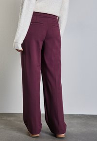 Pantaloni larghi color bordeaux realizzati in tessuto liscio, con tasche posteriori e un fit rilassato, abbinati a un maglione leggero con texture.