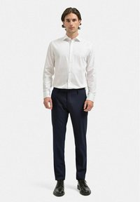 Vit långärmad skjorta med knappar, kombinerad med marinblå raka byxor och svarta skor, stående mot en enkel vit bakgrund.