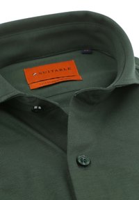 Groene katoenen shirt met een geplooide kraag, voorzien van een oranje label met de tekst "Geschikt" en een zwarte knoop bij de hals. Glad materiaal.
