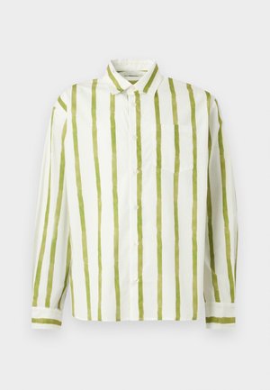 Homme portant une chemise à boutons rayée verticalement verte et blanche, un jean bleu ample, des chaussures noires et des lunettes de soleil foncées, debout devant un fond uni.