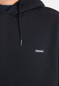 Prim-plan al unui hanorac negru cu o etichetă logo pe partea din față care spune "Dickies" și un șnur atârnând de glugă.