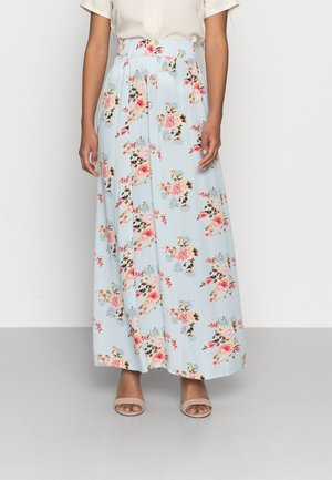 Mujer con falda maxi floral azul claro con flores rosas y marrones, combinada con sandalias beige abiertas y una blusa blanca de manga corta.