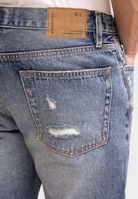 Jeans in denim blu chiaro con dettagli strappati sulle tasche posteriori. Presentano un'etichetta marrone cucita nella vita con "R/R" e "Ispirato dalle Strade Urbane."